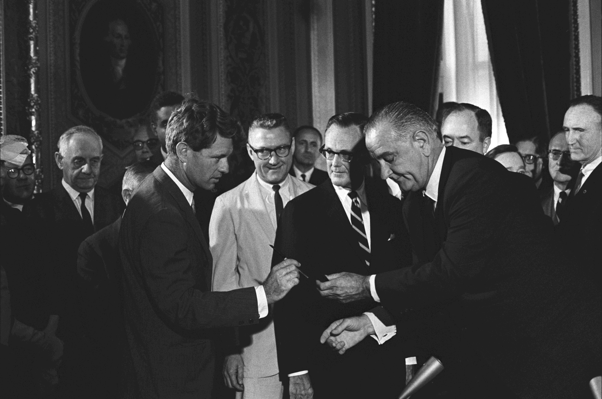 President Lyndon B. Johnson hands a signing pen to Senator Robert F. Kennedy as others look on.