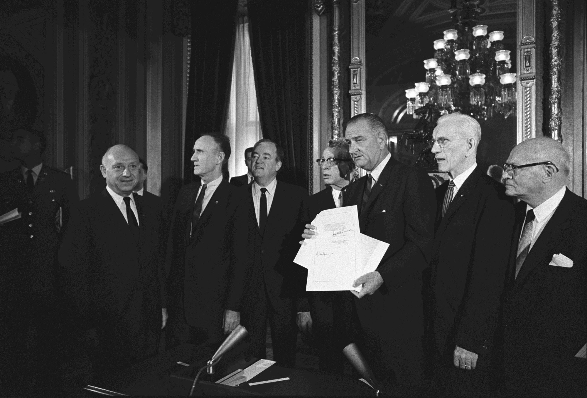 President Lyndon B. Johnson, holding signed copy of the bill, posing for camera with Congressional leaders: (L-R) Sen. Jacob Javits, Sen. Mike Mansfield, V.P. Hubert Humphrey, Sen. Everett Dirksen, Pres. Johnson, Speaker John McCormack, unidentified.
