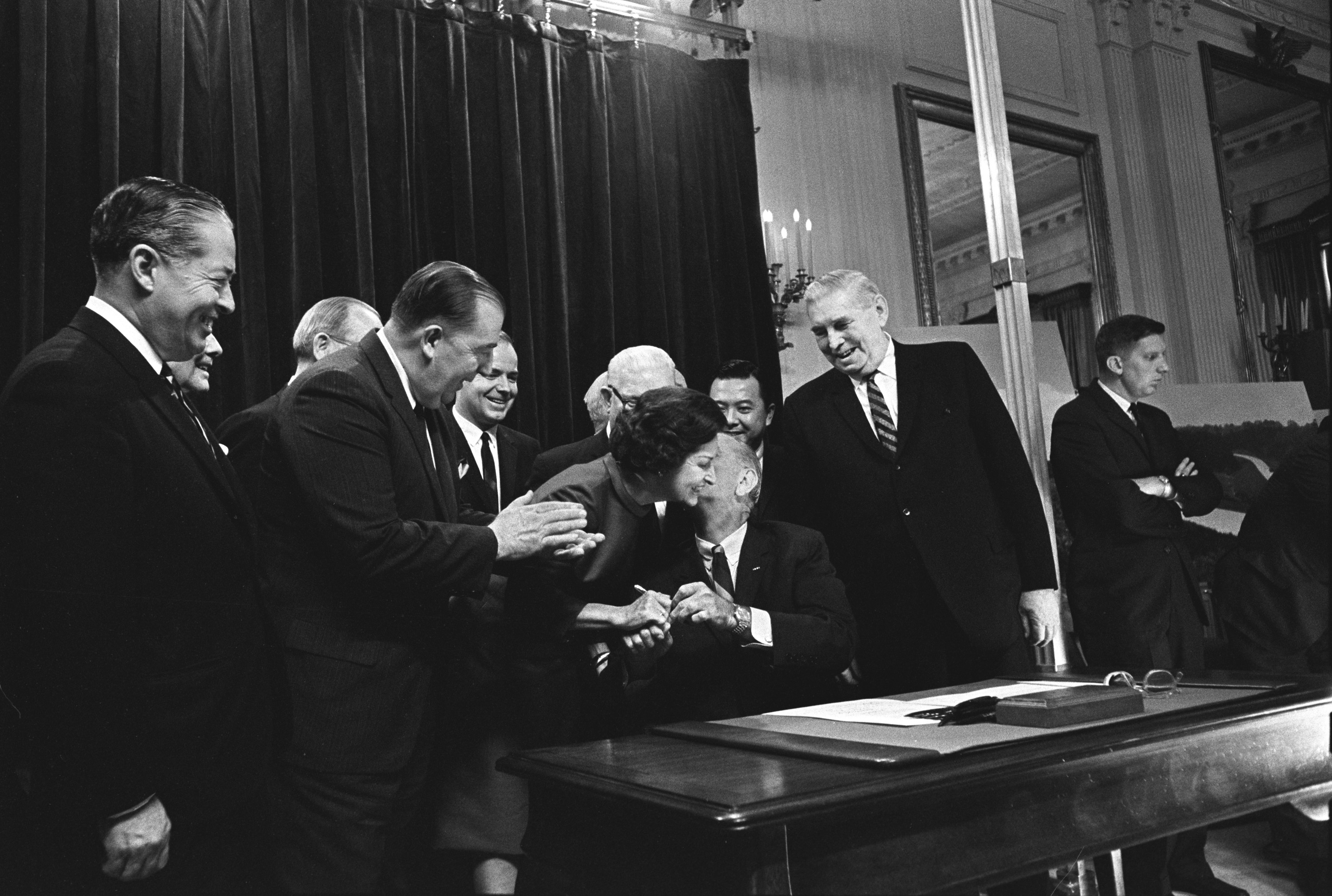 President Lyndon B. Johnson hands a signing pen to Lady Bird Johnson. Others including Sen. Jennings Randolph (applauding), Sen. Daniel Inouye (behind LBJ), and Sen. Pat McNamara (behind & left of LBJ) look on.