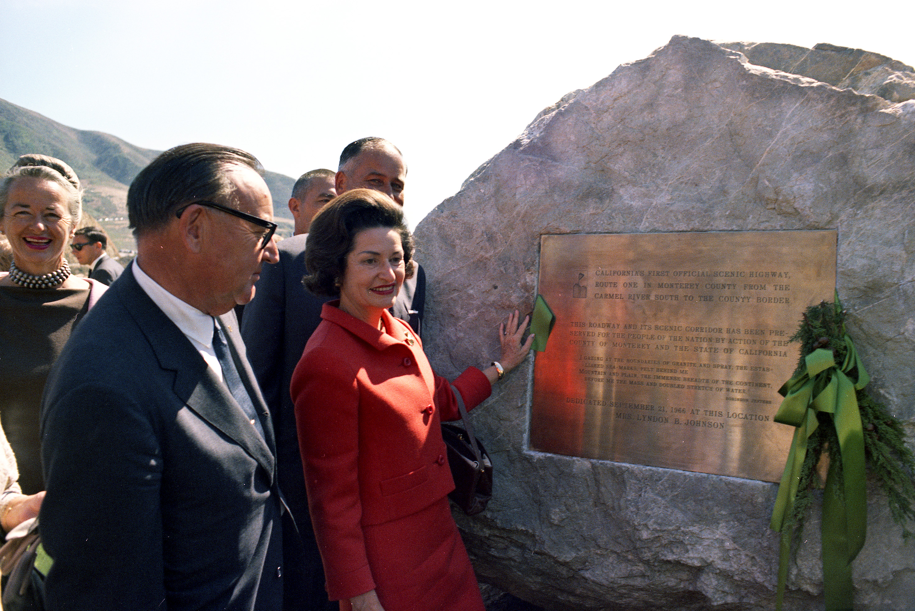 At Bixby Creek Bridge, Lady Bird Johnson poses with a plaque at the dedication ceremony of California's Highway 1 as the country's first scenic highway. California Governor Edmund "Pat" Brown stands at her right.