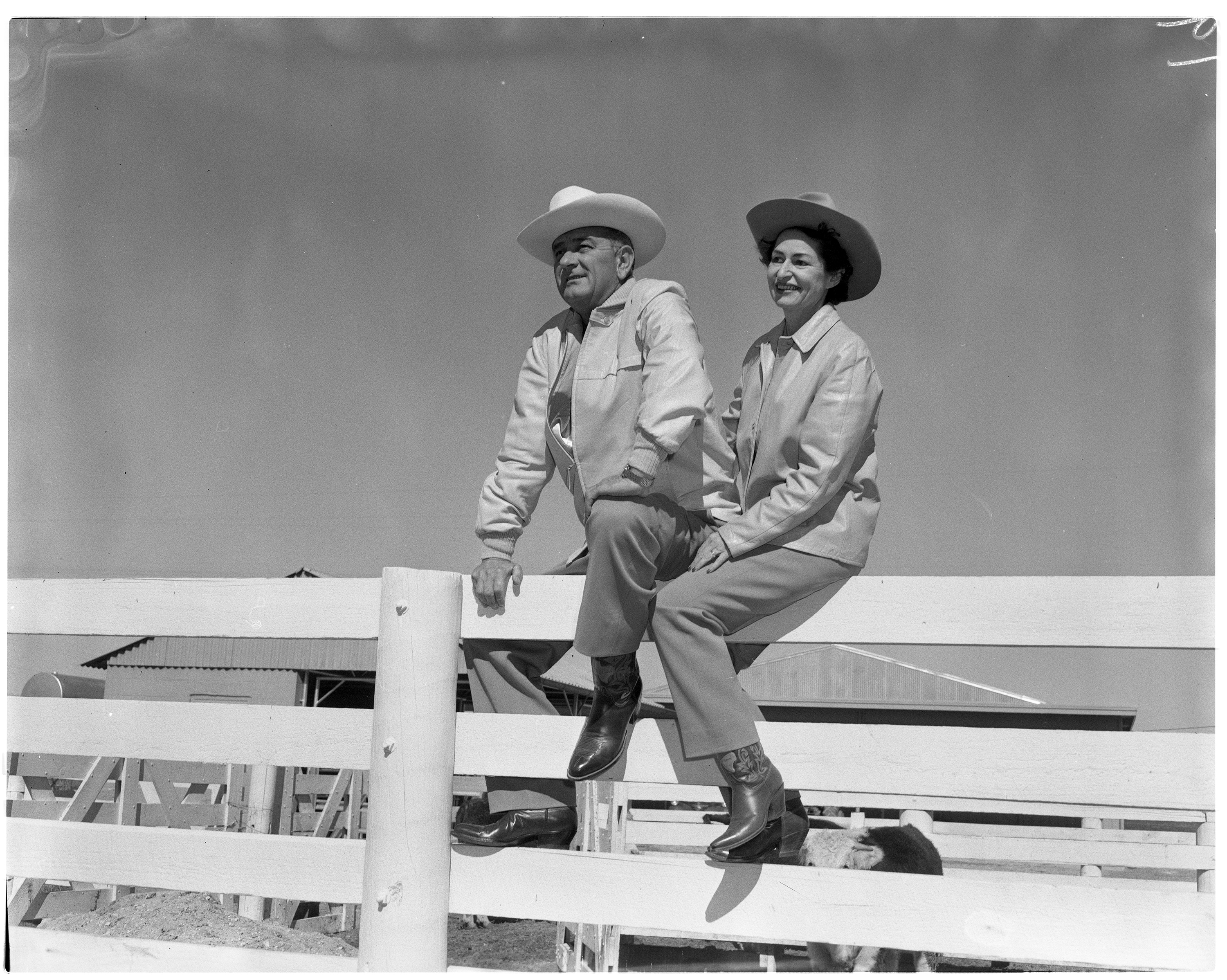 Lyndon B. Johnson and Lady Bird Johnson sit on top of a fence at the LBJ Ranch.
