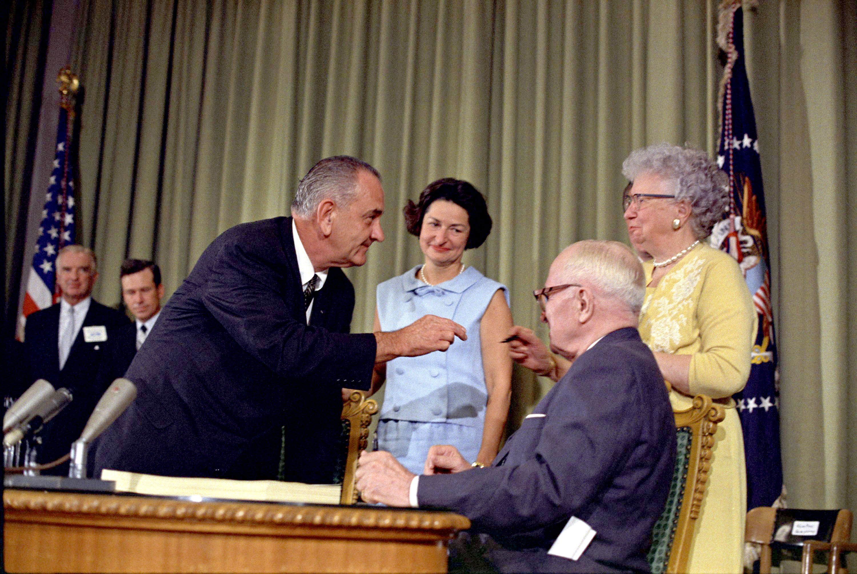 President Lyndon B. Johnson hands a signing pen to Bess Truman as President Harry S. Truman, Lady Bird Johnson and others look on. LBJ Library photo by unknown #34897-16.