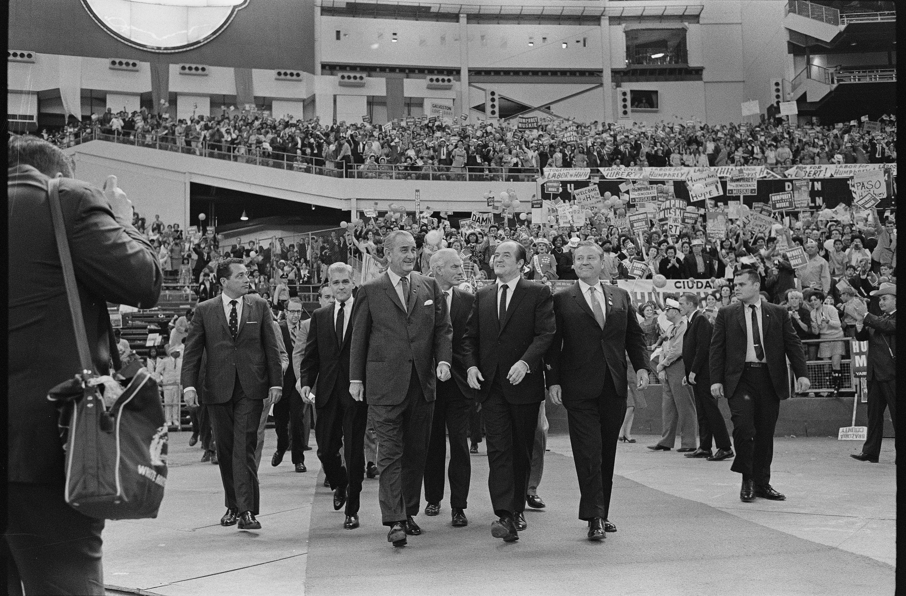 President Lyndon B. Johnson, Vice President Hubert Humphrey, and Sen. Ralph Yarborough walk into the Astrodome as thousands applaud and cheer.