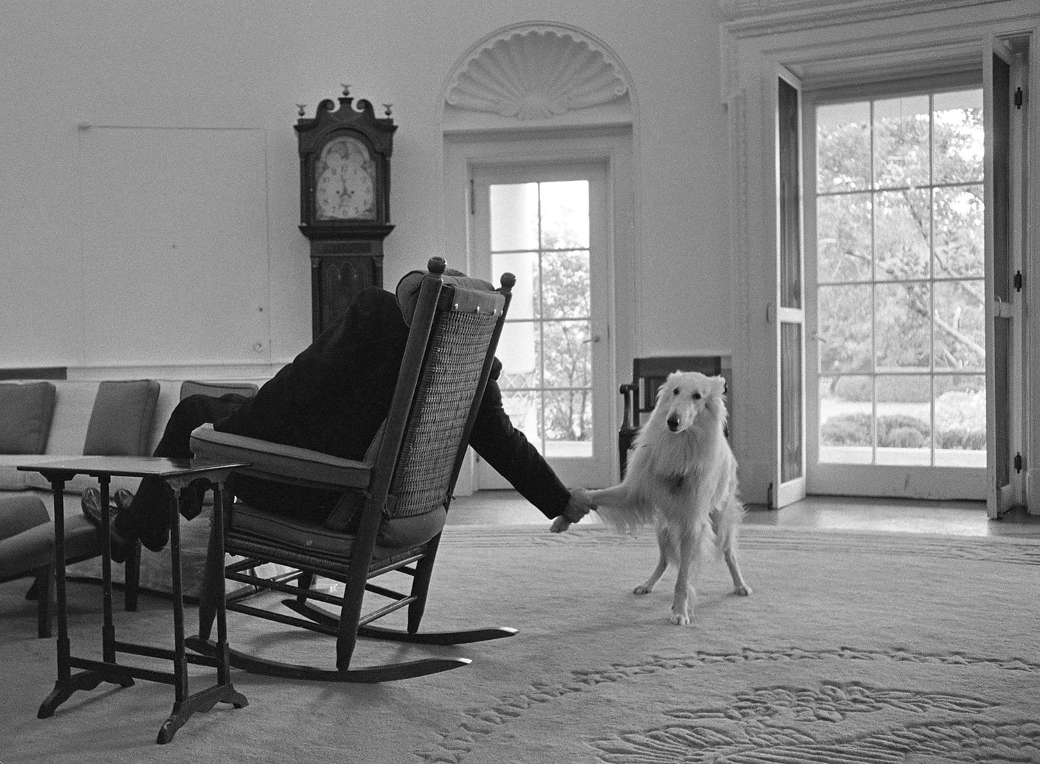 Seen from behind, President Johnson holds Blanco's paw while sitting in his rocking chair in the Oval Office.