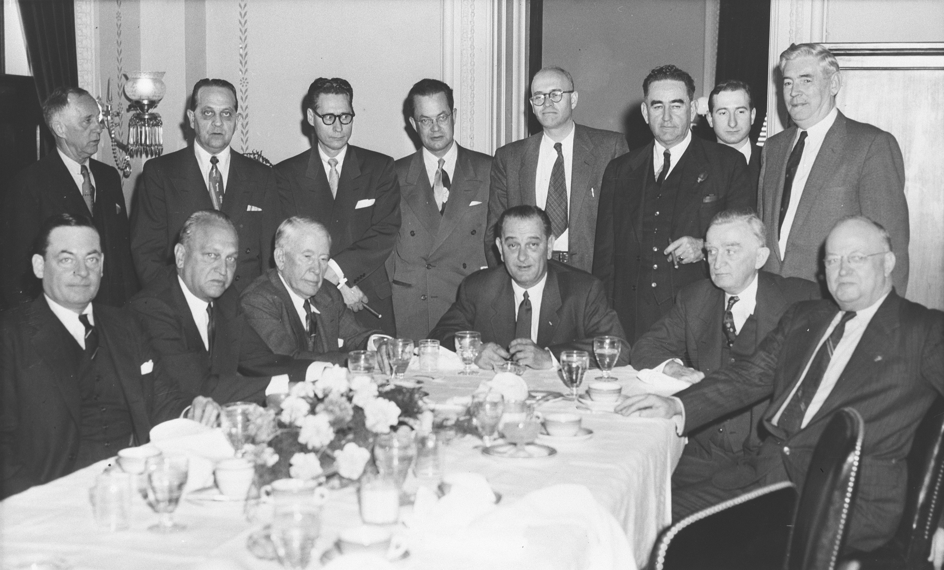 A row of suited men stands behind a row of seated men at a banquet table.