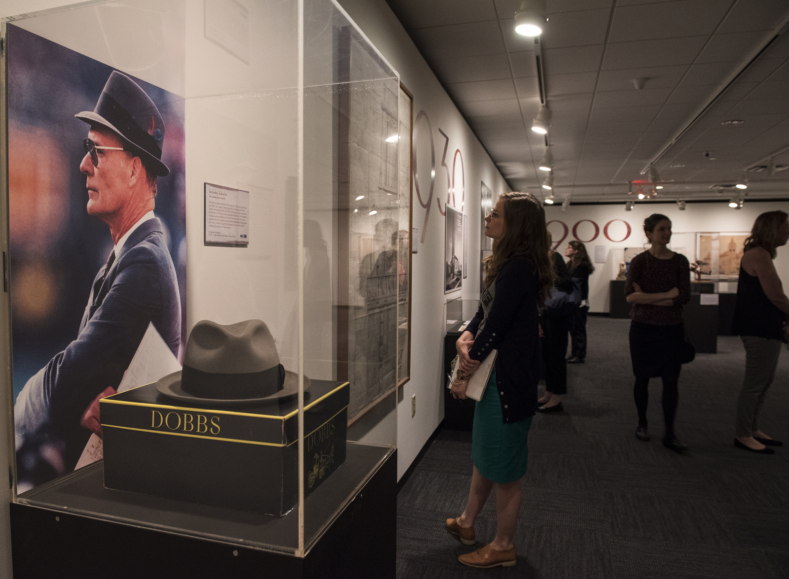 A closeup of a fedora hat worn by Dallas Cowboys Coach Tom Landry, while in the background a visitor examines a painting in the "Deep in the Vaults of Texas" exhibition at the LBJ Library.