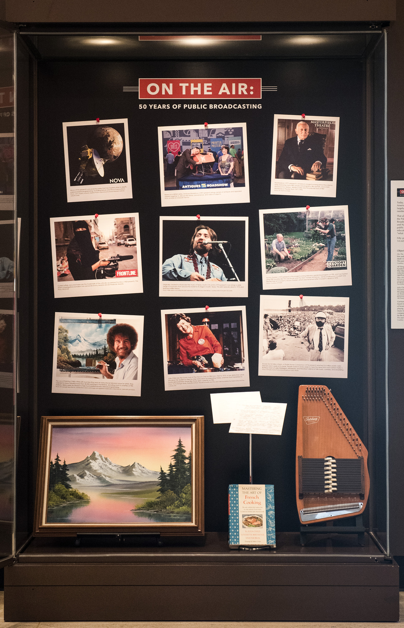 A photo of a glass case displaying several artifacts, including a Bob Ross painting, that was a part of the "On the Air" exhibition at the LBJ Library.