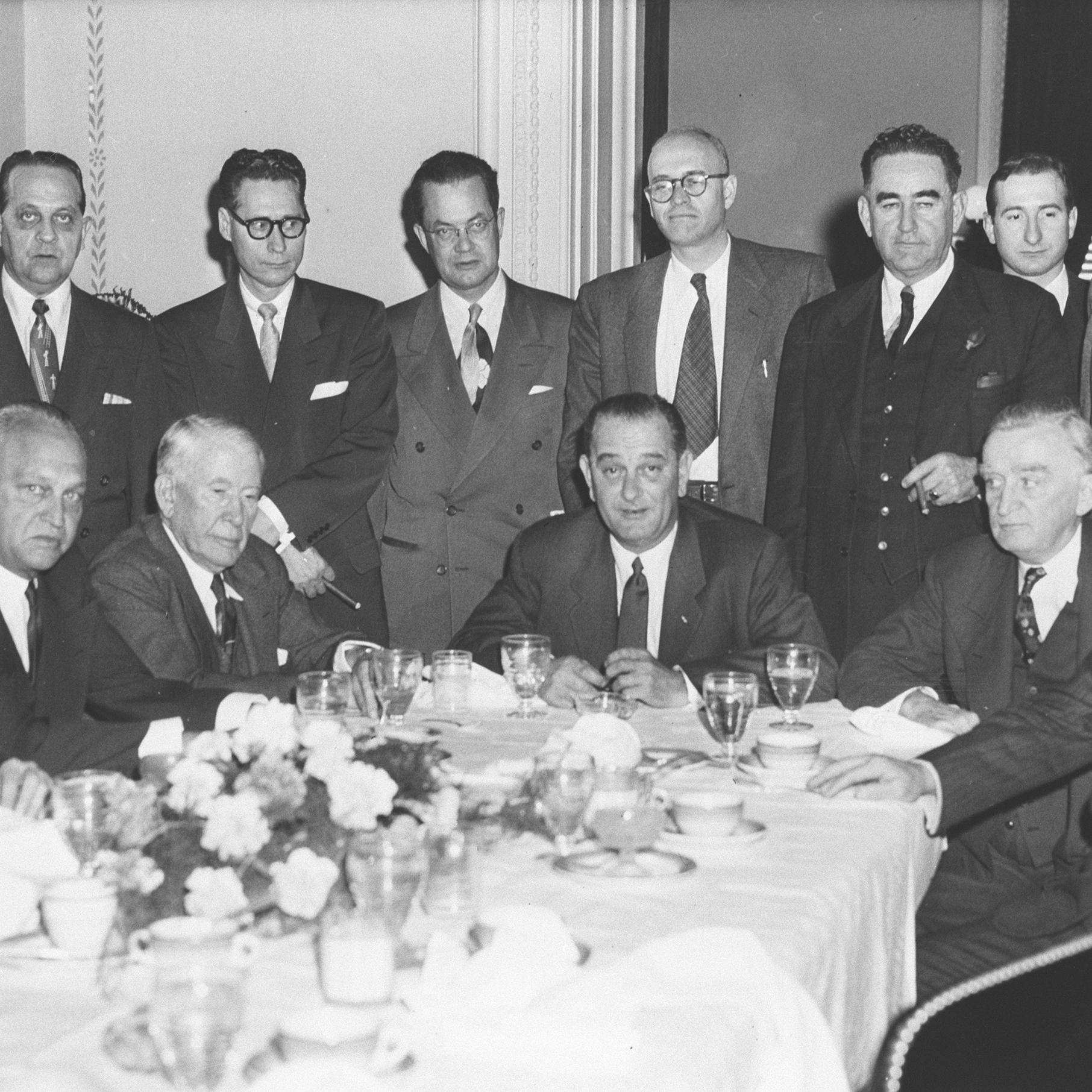 A row of suited men stands behind a row of seated men at a banquet table.