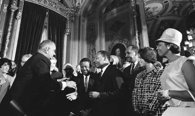 President Lyndon B. Johnson moves to shake hands with Dr. Martin Luther King while others look on.